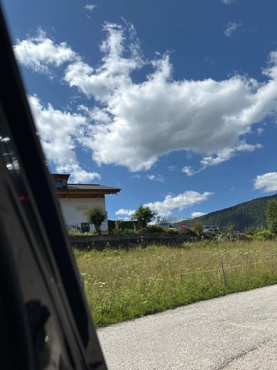 This image captures a view from within a vehicle looking out towards a rural landscape on a sunny day. The foreground is dominated by the dark interior of the vehicle, obscuring a portion of the scene. Beyond the vehicle, a white building with a brown roof and wooden accents is visible, bearing a sign with the text "Gästehaus". A small tree is planted in front of the building.

The midground features a field of tall green grass dotted with white wildflowers, and a low stone wall separates it from a slightly elevated area where several cars are parked. Behind the cars, a cluster of pink flowers adds a splash of color. The background consists of a densely wooded mountain slope under a vibrant blue sky filled with scattered white cumulus clouds. The overall impression is one of a tranquil, picturesque setting in a mountainous region. Based on the sign on the building, the location is identified as Bischofswiesen, Germany.