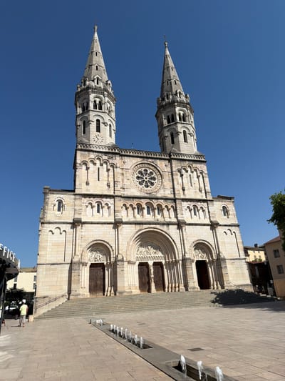 The image shows a large stone church with two tall spires under a clear blue sky. The church has a grand facade with multiple arched doorways, detailed carvings above the entrances, and a large rose window. A wide set of stone stairs leads up to the main entrance. In front of the church, a paved plaza features a long, rectangular water feature with multiple small fountains. To the left of the church, a partial view of a building with the word "OFFICE" on it is visible. A single person, wearing a light yellow t-shirt and shorts, walks away from the camera across the plaza towards the church. To the right of the church, a portion of a building with a terracotta roof and other residential buildings can be seen. The overall impression is a sunny day in a European city. This is the Cathédrale Saint-Vincent de Charnay-lès-Mâcon, located in the city of Charnay-lès-Mâcon, France.