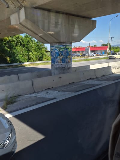 This image captures a view from a vehicle, likely traveling on a highway, looking towards a roadside scene under an overpass. In the foreground, the dark asphalt of the road is visible, with a portion of a gray car on the left side.

The middle ground features a concrete overpass support pillar with posters attached to it. The posters appear to be advertisements for a circus, with images of performers and text that includes "Circo Nation" and "Cuba." Concrete barriers line the edge of the highway. Beyond the barriers, a road with several parked cars is visible, as well as a building with a red roof. A light pole and trees are also present in the background. The sky is clear and blue.

The scene is consistent with the area around the Egnatia Odos highway in Neos Rysio, Greece. The presence of the overpass, highway infrastructure, and the style of the building in the background are characteristic of this region. The overall impression is a sunny day along a major road in a semi-urban or rural area.