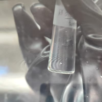 The image shows a close-up of a gloved hand holding a small glass vial. The vial contains a clear liquid and some small particles are visible within it. A label on the vial is partially visible, with text that appears to read "RF 0108" and "400". The background is blurred and appears to be indoors, with some reflections and lighting suggesting an artificial environment. There is no information in the image to suggest the location in Płock, Poland.
