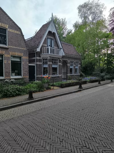 A residential street in Oudorp, Netherlands, features a brick herringbone-patterned road adjacent to a sidewalk with small, dark bollards. A brick house with a gabled roof, white trim, and a balcony is visible. The house also has a small garden bordered by a black metal fence containing plants and flowers. Trees are in the background.