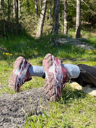 The image displays a close-up view of a person's lower legs and feet, resting outdoors in a natural, sunlit environment. The person wears black pants, light gray ribbed socks, and a pair of maroon and light pink/beige Asics running shoes. The shoes clearly show the "ASICS" logo and the model name "GEL-CUMULUS 16" on the side.
Their feet are propped up on a large, textured gray log or fallen tree trunk, which appears to be firmly settled on the ground. The ground is covered with vibrant green grass, interspersed with small white and yellow wildflowers, indicating a lush natural setting. In the background, numerous tree trunks and green foliage suggest a forest or woodland area. The strong sunlight casts clear shadows, indicating a bright day, likely morning or afternoon. The scene evokes a feeling of relaxation or a break from an outdoor activity in the scenic location of Слизнево, Russia.