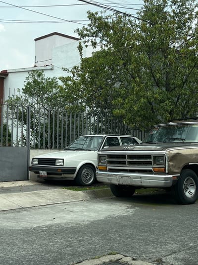 The image depicts a residential street scene in Ciudad López Mateos, Mexico, under an overcast sky. Two vintage vehicles are parked on a paved surface, adjacent to a grey metal gate and a white-fenced property. A white, four-door sedan, possibly a Volkswagen Jetta, is parked on the left, facing right. Its front bumper is slightly raised on a concrete curb, and a license plate reading "MON-342-B" is visible. To its right, a brown pickup truck, identified by the "DODGE" emblem on its hood, is parked, facing left. The front of the truck features a chrome grille and bumper. Behind the vehicles, a mature tree with dense green foliage partially obscures a white building with a dark-capped roof. The environment suggests a quiet, possibly residential area within the city. No people are visible in the image.