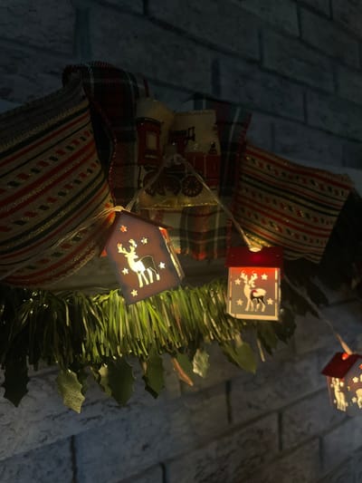 A close-up displays a festive wreath adorned with holiday ribbons and whimsical string lights. The ribbons are patterned with red, white, and black stripes, and feature a train design. The lights consist of miniature wooden houses with reindeer and star cutouts that emit a warm, inviting glow. Green foliage is interspersed among the decorations, adding a natural touch to the festive display.