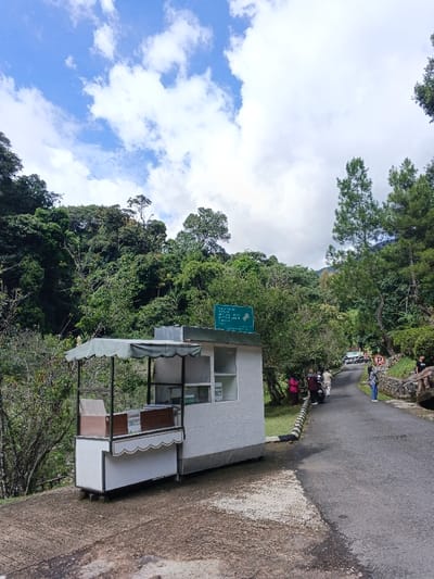 The image captures an outdoor scene at what appears to be a natural tourist attraction or park entrance, likely located in a mountainous or hilly region. The setting is bright and green, suggesting a pleasant daytime environment.

In the foreground, on the left, a small, white vendor stand with a light green wavy awning is visible. It features a wooden counter and shelves, possibly for displaying items for sale. Attached to or placed alongside this stand is a slightly larger, rectangular white building with a flat roof and windows, also with a subtle green trim.

Perched on the roof of this white building is a prominent green sign with white text and an arrow pointing to the right. The text on the sign reads:
- "TEMPAT WISATA" (Tourist Attraction)
- "SATUAN" (Unit, possibly part of a park or management name)
- "BAYAR RETRIBUSI VIA QRIS" (Pay retribution/fee via QRIS)
- "SCAN QR CODE UNTUK" (Scan QR Code for)
- "PEMBAYARAN TIKET" (Ticket Payment)

To the right of the building, an asphalt road curves slightly uphill and away into the distance. The road is bordered on its left by a low curb or barrier with black and white stripes. Further along the road, a small group of people is visible. Some individuals are standing next to what appear to be parked motorcycles or scooters, while others are walking or standing nearby. One person is seen further down the road, near a stone wall. The people are casually dressed, indicating a leisurely outing.

The background is dominated by lush, dense green forest and trees covering the hills, extending high up towards the horizon. On the far right, several tall, slender pine-like trees stand out against the backdrop. The sky above is a clear blue with scattered white cumulus clouds, indicating fair weather. The overall impression is one of a serene natural environment with infrastructure for visitors.