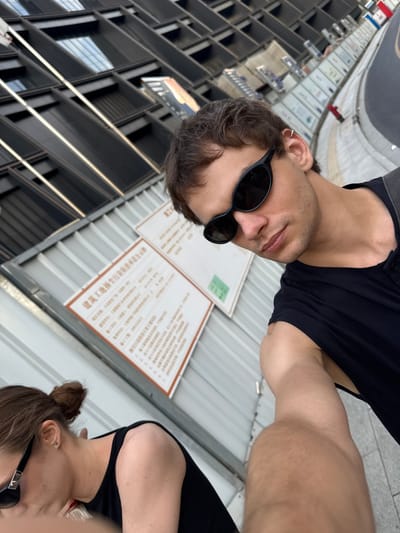 This rotated image captures a selfie-style view of two individuals outdoors in Putuo District, China, under bright daylight conditions. In the foreground, positioned towards the left, is a man with dark hair and light stubble, wearing black sunglasses and a black sleeveless or short-sleeved top. A silver hoop earring is visible in his left ear. His right arm, muscular and light-skinned, extends across the upper right portion of the frame.

Partially visible on the right side, slightly behind the man, is a woman. Her head, neck, and part of her shoulder are seen. She has brown hair tied in a bun, wears dark sunglasses, and a black top.

The background features distinct urban elements. Behind the subjects, a light gray corrugated metal fence or wall stretches horizontally. Affixed to this wall are at least two white rectangular signs covered with Chinese characters, suggesting official notices or information. To the left and top of the frame, a modern building with a dark, grid-like architectural facade is visible, reflecting sunlight. To the far right, a gray paved surface, likely a sidewalk or road, completes the scene. The overall ambiance is casual, with both subjects wearing sunglasses, obscuring their expressions.