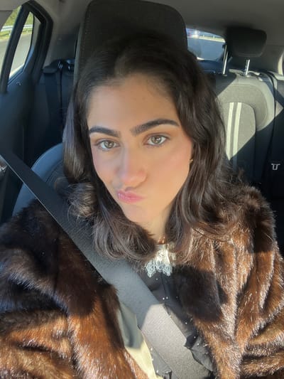 An individual is observed seated in the front passenger seat of a vehicle, located in Caminha, Portugal. The subject, an adult female with dark, wavy hair and brown eyes, is depicted looking directly at the camera with her lips pursed. She is wearing a dark brown, fur-like coat over a black garment featuring a white lace collar. A dark gray seatbelt diagonally crosses her chest. The vehicle's interior shows dark upholstered rear seats, partially illuminated by direct sunlight which casts shadows and highlights on the subject and the interior. A blurred background visible through the vehicle's rear window indicates an outdoor roadside environment with green foliage.