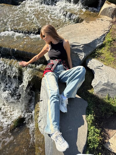 A female individual is seated on a large, flat gray rock next to an artificial multi-tiered waterfall in Veshki, Russia. She is wearing a black sleeveless tank top, loose-fitting light blue denim jeans with faded vertical stripes down the outer seams, and white and light gray athletic sneakers. A dark gray vest or cincher with red floral embroidery and structured vertical elements is worn over her top. Dark sunglasses are on her face, and her light brown hair extends over her shoulders. Her left arm is extended, with her hand touching the surface of the flowing water. The immediate environment consists of large, irregular gray rocks forming the waterfall and its banks, with green ground cover vegetation visible on the right side and in the background. The scene is brightly illuminated by direct sunlight, casting distinct shadows.