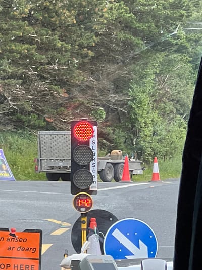 In Creeslough, Ireland, a temporary traffic light displays a red signal, indicating a stop. The traffic light is mounted on a pole and has three signal heads, with the top one illuminated red. Below the red light, two dark signal heads are visible. A digital timer is positioned below the traffic light, showing "1:42".
In the background, a small trailer with a cage-like structure is parked. Two traffic cones, one solid orange and the other orange and white striped, are placed on the road. Further back, a dense line of trees and foliage forms the backdrop.
In the foreground, an orange traffic sign with white lettering reads "n anseo ar dearg" and "OP HERE". To the right of this sign, a blue circular road sign with a white arrow pointing downwards and to the left is visible. Several people, mostly obscured by the vehicle the photo is taken from, are implied to be present, as suggested by the context of a traffic control situation. The overall scene depicts a temporary traffic management setup, likely due to road work or an event in Creeslough.