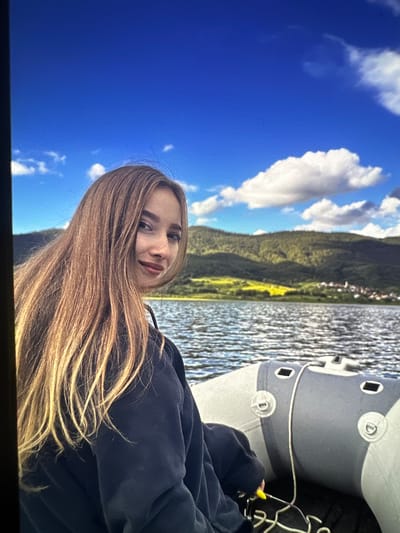 A young woman with long, blonde hair is seated in a boat on a body of water, looking towards the camera with a slight smile. She is wearing a dark blue sweatshirt. In the background, rolling hills covered in green vegetation and a village with red-tiled roofs are visible under a bright blue sky with scattered clouds. The boat appears to be an inflatable dinghy with a gray and white exterior. The water is rippled, suggesting a gentle breeze. The scene evokes a sense of leisure and outdoor activity. While the image shows a scenic landscape with a village, it is not possible to definitively identify the specific location within the city of Dragoman, Bulgaria, based solely on the visual information provided.