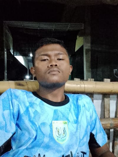 A young man with dark skin and short black hair is looking directly at the camera. He has a faint beard and mustache. He is wearing a light blue t-shirt with a black collar and a pattern of light blue shapes. On the left side of his chest, there is a shield-shaped logo with green and yellow accents and some writing. He is sitting on a bench made of bamboo poles. The background is dark and appears to be outdoors at night, possibly a covered porch or patio with some lights visible in the distance. The overall scene is candid and informal.