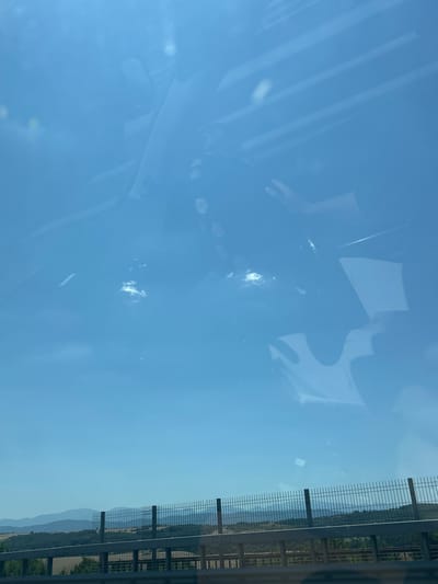 The image shows a view from inside a vehicle, looking out through the windshield. The sky is bright blue with some faint reflections and a few wispy clouds. In the foreground, a metal fence runs horizontally across the frame, separating the road from the landscape beyond. Beyond the fence, rolling hills covered in greenery and some dry fields stretch into the distance, with a hazy mountain range visible on the horizon. The scene appears to be set on a sunny day, possibly during the warmer months. The location is identified as Vakarel, Bulgaria.