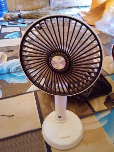 This media file captures a close-up, eye-level view of a small, white desk fan positioned on a patterned surface. The fan's blades are dark, arranged radially around a central metallic hub, and are partially obscured by a dark grille. The fan's body and base are white, with a small circular logo or symbol visible on the neck of the fan. The base is circular and slightly wider than the fan's head.
The background of the image suggests an indoor setting, possibly a room or a covered outdoor area. The patterned surface beneath the fan is a mosaic of beige, brown, and blue squares, with a large blue floral design visible on the left. In the upper left corner, a broom with a bundle of dry straw tied with a black band is visible, lying horizontally. To the right of the fan, a crumpled dark plastic bag and some beige material are present. The overall lighting appears to be natural, suggesting daytime. No people are visible in the image. The scene evokes a sense of everyday domesticity. The visible text on the fan is a small circular logo, possibly an initialism or brand mark. The location context is Dundaye, Nigeria.