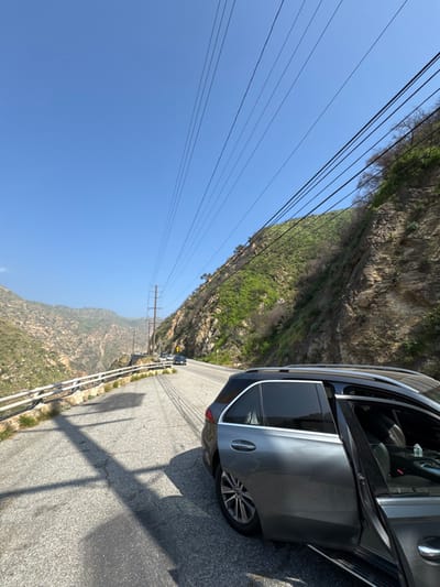 The image displays a scene along a paved mountain road in the Unincorporated Santa Monica Mountains, United States of America, under a clear blue sky. In the immediate foreground, a dark gray SUV, likely a Mercedes-Benz GLE-Class, is parked on the right shoulder of the road, with its front passenger door ajar. A white water bottle is visible inside the open door. The vehicle casts long, distinct shadows across the asphalt to the left. The two-lane road curves leftward and ascends into the distance, where several other vehicles are observed. A white metal guardrail lines the left side of the road, overlooking a steep, rocky canyon. To the right of the road, a steep hillside covered with rocky outcrops and green vegetation rises sharply. Utility lines extend across the upper portion of the frame, supported by at least one visible utility pole in the midground. No individuals are visible within the scene.