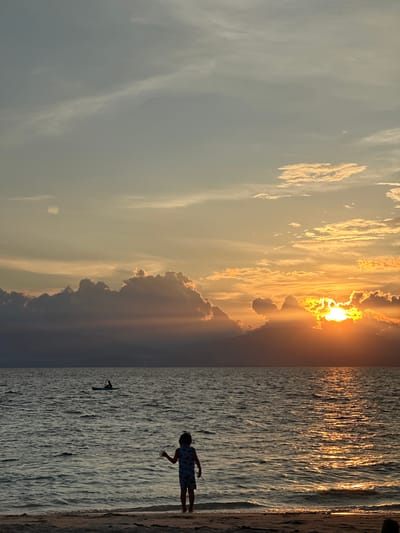 The image depicts a serene sunset scene on a beach in Saavedra, Philippines. The sky is filled with a gradient of colors, from pale yellow and orange near the horizon to a soft blue higher up, with scattered clouds. The sun, partially obscured by clouds and mountains in the distance, casts a brilliant golden glow on the rippling water.
In the foreground, a child stands at the water's edge on a sandy beach, facing the ocean and raising one hand as if waving. The child is silhouetted against the sunset. Further out in the ocean, a lone figure can be seen paddling a small boat, possibly a kayak or paddleboard. The water appears calm, with gentle waves lapping at the shore. The overall atmosphere is peaceful and tranquil, capturing a moment of natural beauty and quietude.