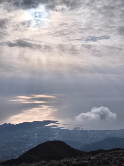 This panoramic image, captured from a high elevation in Mali Mikulići, Montenegro, presents a vast coastal landscape under a heavily clouded sky. The sun, a bright white disc, is partially obscured by dense grey and white clouds in the upper left, casting diffuse light and faint crepuscular rays downward into the atmosphere. Beneath the clouds, the sea reflects elongated streaks of sunlight, indicating a surface interaction with direct light. A prominent coastline features dark, undulating hills and peninsulas, framing a sprawling urban area with discernible buildings and infrastructure, including a significant port facility with docks and cranes on the right side of the main bay. A singular white cumulus cloud hovers independently above the cityscape in the mid-right. The immediate foreground shows the dark, rugged silhouette of a mountain ridge, from which the photograph appears to have been taken. No individuals are present in the scene.