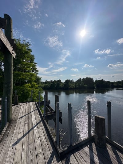 The image shows a wooden dock extending over a body of water, likely a lake or river. The sun is high in the sky, creating bright glare and reflections on the water surface. Trees line the opposite bank of the water. The dock is constructed of wooden planks, with wooden posts and metal supports visible. The sky is blue with scattered white clouds. The scene appears to be set in a natural environment, potentially a park or recreational area.
While the image does not allow for definitive identification, based on similar landscapes in the Tampa area of the United States, it could be a lake in a park, or a river access point. More precise identification would require additional information or visual cues.
