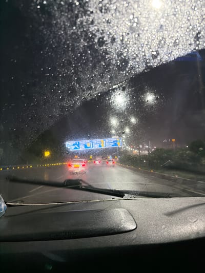The image depicts a nighttime view from inside a vehicle, looking through a rain-streaked windshield. The scene is set on a highway, likely in or near Manchirevula, India, given the context. The road ahead is wet, reflecting the lights of passing cars and overhead street lamps. A prominent blue highway sign with white text and symbols is visible, indicating upcoming directions. Several vehicles, identifiable by their red taillights, are on the road. The foreground is dominated by the car's dashboard and the windshield wipers. The overall atmosphere is dark and wet, characteristic of driving in rainy conditions at night.