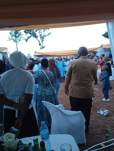 The image depicts an outdoor social gathering, likely a celebratory event such as a wedding reception or party, taking place under large tents in Shimbi Mashariki, Tanzania. The scene is bustling with numerous people, indicating a lively atmosphere.
In the immediate foreground, the back of a person dressed in a white uniform and apron, presumably a server or caterer, is visible. Adjacent to them, a table covered with a white tablecloth holds a bouquet of white roses, a plastic water bottle, and some empty beer bottles. A white chair with a fitted cover stands beside the table. Crutches are also visible on the ground nearby.
The mid-ground features several attendees, including a woman in a blue patterned dress with her back to the camera, and a man in a tan jacket and dark pants, also facing away. Further into the background, a crowd of people is gathered, many smiling and interacting. A woman in a bright blue dress is prominently featured in the center-left background, actively dancing with her arms raised, holding a cell phone, suggesting a festive mood with music. A small child in a blue outfit stands to the right. Many women are dressed in various shades of blue, indicating a possible theme or coordination.
The event appears to be set on an unpaved ground, possibly dirt or sparse grass, with some discarded items like crumpled paper and bottles visible. The bright sky in the background, with two silhouetted trees, suggests it is daytime, possibly late afternoon, with soft, ambient lighting. The overall impression is one of a vibrant community celebration. No visible text is discernable in the image.