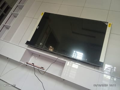 In Nawfia, Nigeria, a wide, flat-screen television is laid face-down on a white, multi-tiered cabinet against a tiled wall. The television, likely new, has protective plastic film and yellow warning stickers on its edges. A white electrical cord trails from the television to a power strip placed on a lower shelf of the cabinet. The cabinet itself is modern and sleek, with clean white surfaces and open shelving. In the background, a partially visible fan and a window with blinds can be seen. The image is timestamped with "REDMI A3" and "06/19/2026 18:11".