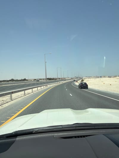 The image captures a multi-lane highway scene from the perspective of a vehicle, looking forward and slightly to the left. The time of day is clear and sunny, indicated by the bright, cloudless blue sky.
In the foreground, the right side of the image reveals part of the interior and exterior of the vehicle from which the photo was taken, including a white hood or fender and a black windshield pillar and dashboard section. The highway stretches into the distance, featuring dark asphalt and clear road markings, including solid yellow lines along the shoulders and white dashed lines separating the lanes.
In the mid-ground, a black car, possibly an SUV or hatchback, is visible driving in the leftmost lane. Its rear license plate, though not entirely legible, shows Arabic script and numbers. Further along the highway, other vehicles appear as small silhouettes. The road is flanked by grey guardrails and wide shoulders of light-colored, sandy ground, suggesting an arid or desert environment. Tall streetlights with horizontal arms line both sides of the highway, extending far into the background.
The background shows a flat horizon with faint indications of distant structures or vegetation in the sandy landscape. The overall scene depicts a modern, well-maintained highway cutting through an open, arid region, consistent with the geographical context of Umm Salal Muhammad, Qatar.