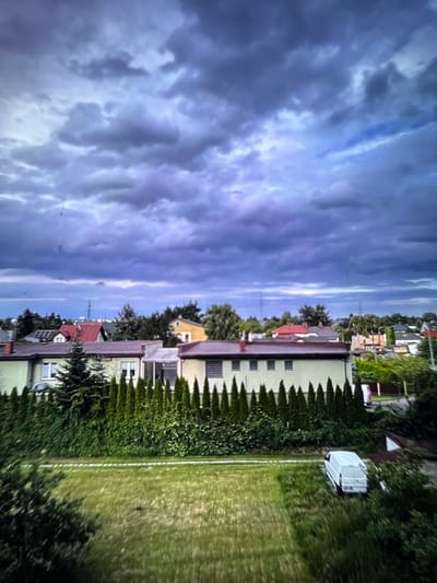 The image shows a residential neighborhood with houses and lush green trees under a dramatic, cloudy sky. The sky is a deep purple and blue, with streaks of white clouds suggesting an approaching storm or recent rain. The houses are varied in color and style, with some featuring red roofs and others a pale green facade. They are lined up along a street, with dense hedges and trees providing a verdant barrier between properties. In the background, more houses and trees are visible, suggesting a densely populated suburban area.
To the right of the houses, a well-maintained green lawn stretches out, bordered by a white line, possibly indicating a sports court or pathway. A white, boxy structure, possibly a storage unit or a small shed, sits on the lawn. The lighting suggests it might be late afternoon or early evening, as the sky is dark but there's still enough light to see the details of the scene. The overall mood is somewhat ominous due to the dark clouds, but the vibrant green of the lawn and trees adds a sense of life and natural beauty. The image appears to be taken from a slightly elevated position, offering a panoramic view of the neighborhood.