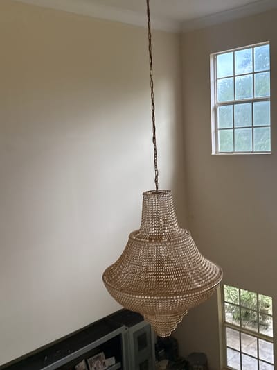 The image shows an indoor scene with a focus on a large, beaded chandelier hanging from the ceiling. The chandelier is off-white or light beige in color and has a complex, multi-tiered design. It hangs from a long, bronze-colored chain that stretches across the image.
Two windows are visible in the background. One window, located on the left, has a grid pattern and appears to be letting in daylight. The other window, located to the right of the chandelier, is also gridded and shows greenery outside, suggesting trees or foliage.
To the right of the chandelier, a dark gray or black cabinet is visible, with items displayed inside. The walls are painted in a neutral, light beige color. The scene is well-lit, suggesting natural light coming from the windows.
The image location is Davie, United States of America.
