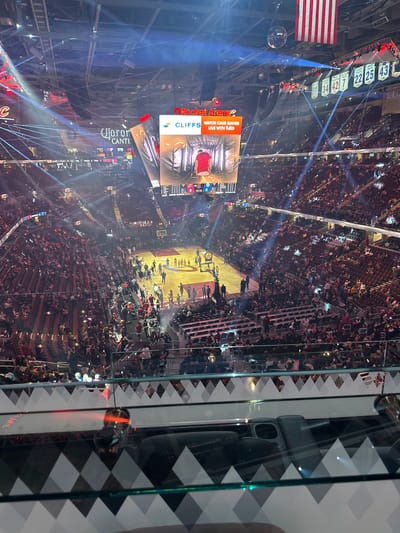 The image shows a basketball game inside Rocket Mortgage FieldHouse in Cleveland, Ohio. An American flag and retired jerseys are visible near the ceiling. The court is lit up, with players visible on the court.  A large crowd is present in the stands, and a digital display screen shows game information and advertisements. The image is taken from an elevated position behind glass, possibly a suite.