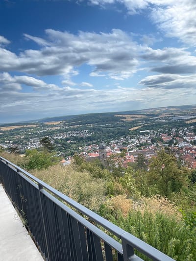 The image presents a scenic vista of Homberg (Efze), Germany, captured from an elevated viewpoint. In the foreground, a modern grey metal railing and a concrete walkway suggest a viewing platform or balcony. Beyond the railing, lush green vegetation, including trees and shrubs, partially obscures the view of the town below.

The town of Homberg (Efze) spreads out across the middle ground, characterized by a dense cluster of red-tiled roofs and white-walled buildings. A prominent church tower, appearing to be made of stone, stands out within the urban landscape. The town is nestled amidst rolling hills and fields, with patches of cultivated land and darker green forests extending towards the horizon.

The sky above is a bright blue, dotted with dramatic, fluffy white and grey clouds. The overall impression is one of a clear, possibly late afternoon, day with good visibility. The image is a landscape shot, focusing on the town and its surroundings, with no people or discernible activity visible. The composition suggests the photograph was taken from a high vantage point overlooking the town.