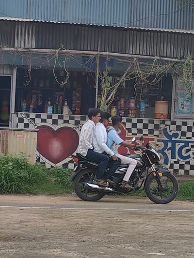 Three young men are riding a motorcycle on a paved road in Malkapur, India. The man in the front is driving, and the other two are passengers. The motorcycle is black and has a red stripe on the side. The men are wearing casual clothing. The background shows a building with a corrugated metal roof and a checkered pattern on the wall, with a large red heart painted on it. There are plants growing on the building and a shop with shelves of items visible inside. The road is dirt and gravel, and there is grass on the side. The sky is overcast.