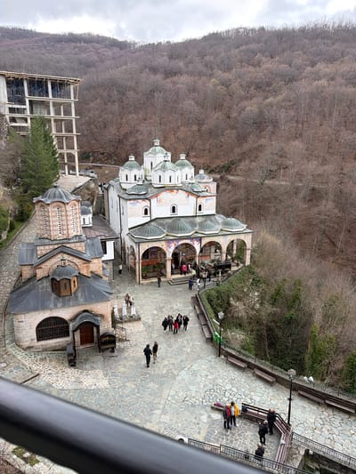 The image shows a top-down view of the St. Joakim Osogovski Monastery complex, located near Kriva Palanka, North Macedonia. The monastery is comprised of multiple buildings with distinct architectural features, including domed roofs, archways, and stone facades. Some buildings display decorative elements and frescoes. A large, modern concrete structure is visible adjacent to some older buildings, possibly indicating recent construction or renovation.

Several people are present in the image, primarily on a paved stone courtyard. Some people are walking, while others are sitting on benches along the edge of the courtyard.

The monastery complex is situated on a hillside. A dense forest with bare trees covers the slope behind the monastery. The sky is overcast.