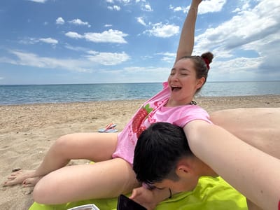 The image depicts a scene on a beach in Νέα Φλογητά, Greece. A woman in a pink t-shirt with a strawberry graphic is sitting with her legs extended, one arm raised in the air. Her mouth is open in a wide smile, and her eyes are partially closed. Her hair is tied up in a bun with a red hair tie. A man lies beneath her, face down on a green towel, with his head resting on the sand. He appears to be holding a phone. Flip-flops are visible in the background, along with the sea and a cloudy sky. The beach consists of sand.