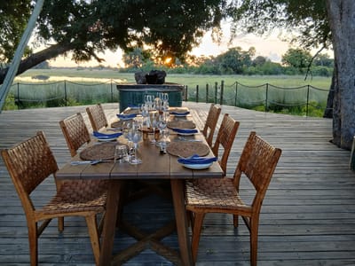 The image depicts a set outdoor dining area on a wooden deck overlooking a natural landscape, possibly near a body of water. The setting is likely in a safari or wildlife area of Nwanaeanoka, Botswana, based on the visible vegetation and setting sun.
The dining table is set for eight people, with each place setting featuring a plate, woven placemat, silverware, glassware (including wine glasses), and a blue napkin. The chairs are made of wood and have woven backs and seats.
In the background, there is a structure that appears to be a green fire pit or decorative element. Beyond that, there is a view of tall grasses, trees, and a water body reflecting the sunlight. A tree provides shade over the dining area. A rope fence serves as a barrier. No people are visible in the image.