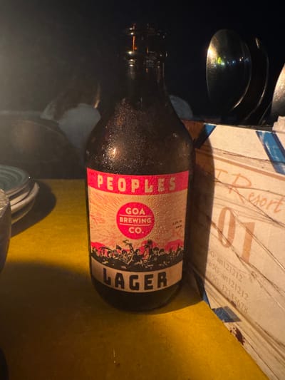 The image forensically displays a dark brown glass bottle, coated in condensation, resting on a bright yellow tabletop. The bottle's label is predominantly pink and light beige, featuring "PEOPLES" in red at the top. Below this, a red circle with radial lines contains "GOA BREWING CO.", and to its right, "4.5% ABV" and "330 ml" are visible. The lower portion of the label depicts a silhouette of a crowd holding flags, above which "LAGER" is printed in white capital letters on a dark background strip.

To the immediate left of the bottle, a stack of white ceramic plates is partially visible. To the right, a light-colored, possibly wooden, stand or holder displays handwritten text including "Resort", the number "01", and alphanumeric codes such as "SG: an121212" and "SG: 12345678". Beyond this stand, metallic reflections, consistent with cutlery, are observed. The background is dimly lit and largely blurred, showing indistinct shapes that suggest other individuals or objects within the environment. The scene is consistent with a resort dining or bar area during evening hours in Kolamb, Goa, India, as evidenced by the "GOA BREWING CO." product and the "Resort" signage.