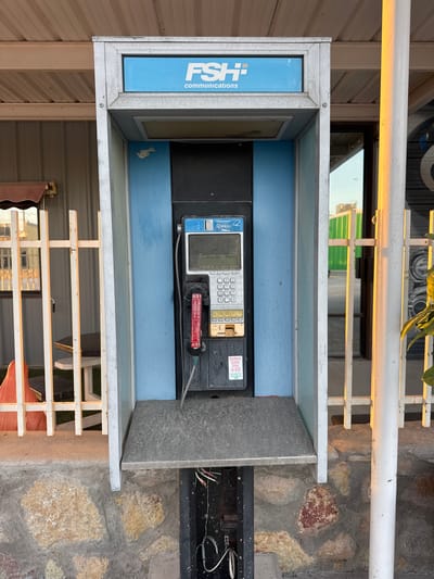 A public payphone booth, likely located in Sunland Park, New Mexico, United States of America, stands in front of a building with a corrugated metal exterior. The booth itself is constructed with a galvanized metal frame and blue panels. At the top of the booth, a sign displays "FSH Communications" in blue lettering on a white background. Inside the booth, a payphone unit is mounted. The phone features a dark gray casing with a digital display screen and a keypad. A black handset is attached by a coiled cord. Below the keypad, there is a coin slot and a card reader. A sticker on the lower part of the phone reads "Collect Calls Dial *89". The payphone booth sits on a stone foundation, and the area around it appears to be outdoors. A white metal fence runs along the side of the booth, and in the background, a building with green siding and reflections of the sky can be seen. The lighting suggests it may be late afternoon, with warm sunlight illuminating the scene.