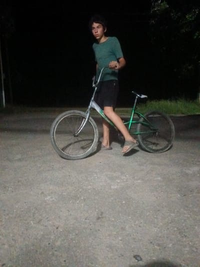 An underexposed, medium shot captures a young person standing next to a bicycle on a paved surface at night. The individual, appearing to be of male gender and of juvenile age, is wearing a teal t-shirt and black shorts. They are barefoot, with one foot visible wearing a sandal. The bicycle, a BMX-style bike with a green frame and silver handlebars, is positioned centrally in the frame. The wheels are prominent, with the front wheel larger than the rear. The background is largely dark, with hints of greenery and trees suggesting an outdoor environment, possibly a residential area or park. The paved surface appears to be asphalt or concrete, with some texture visible. No specific landmarks or signs indicating the location in Leontyevskoye, Russia are present in the image.