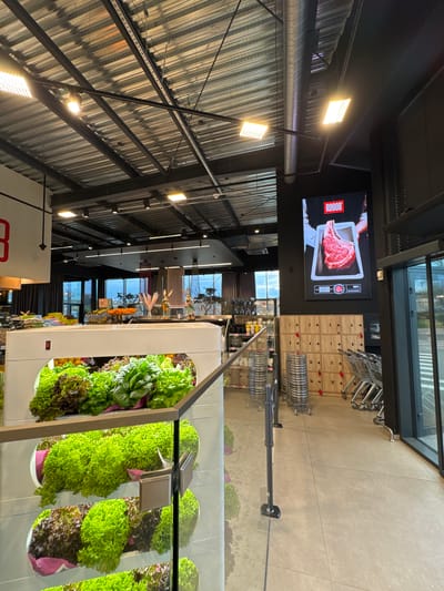 The image depicts the interior of a ROGOB supermarket, likely located in Ciorescu, Moldova. The foreground shows a display of fresh produce, including lettuce arranged in vertical rows. Above, a digital screen shows an advertisement for ROGOB meat products, specifically a raw beef steak. Adjacent to the screen, there are wooden lockers and stacked shopping carts. The store has a high ceiling with exposed metal beams and industrial lighting. Further back, other product aisles and displays can be seen. The overall impression is of a modern and well-lit grocery store environment.