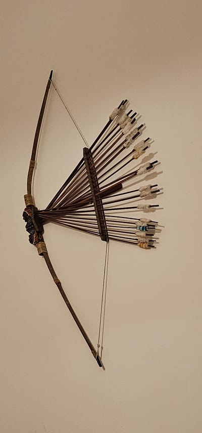An ornate, antique bow and arrow set is mounted on a plain, light-colored wall. The bow, made of dark wood, is curved, with a string tautly stretched across it. At the center of the bow, where the arrows are fletched, is a dark, carved ornament that appears to be a human face. The arrows are fletched with white material, and some have hints of color, such as blue and orange, visible near the nock. The arrows are arranged in a fan-like pattern, held together by a darker wooden structure attached to the bow. The overall impression is of a decorative or historical item displayed as art. There is no indication of a specific location in Dogato, Italy, within the image itself.