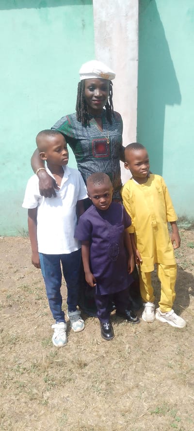 This vertical, full-body shot captures an adult woman and three young boys posing outdoors. The woman, positioned centrally, has dark skin, medium-length dreadlocks or braids, and wears a white beret. Her attire includes a V-neck patterned dress featuring green, black, and red geometric designs, a simple gold-colored necklace, and a gold-toned bracelet on her right wrist. She has her right arm around the boy on her left and offers a slight smile to the camera.
To her left, a young boy with short hair wears a white short-sleeved polo shirt, dark blue jeans, and light-colored sneakers with velcro straps, looking slightly away from the camera. In front of the woman, a younger boy in a dark purple traditional tunic and trousers, paired with black shiny dress shoes, looks directly at the camera with a neutral expression. To the woman's right, another young boy in a bright yellow traditional tunic and trousers, and light-colored sneakers, also looks slightly away.
The background features a light teal or mint green painted wall, with a lighter vertical pillar or corner on the right side. Strong, distinct shadows of the individuals are cast on the wall, indicating bright, direct sunlight during the daytime. The ground beneath them is covered with dry, sparse brown-green grass and dirt. The group appears to be casually posing for a photograph. No visible text is present in the image.