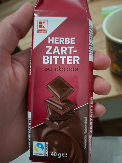 In Ilava, Slovakia, a person is holding a box of K Classic HERBE ZARTBITTER Schokolade. The box is primarily red and white, with the K Classic logo prominently displayed. The text "HERBE ZARTBITTER Schokolade" is printed vertically on the side of the box in white. The front of the box features an image of stacked dark chocolate squares. Below the chocolate image, there are two seals: a Fairtrade logo and a "KAKAO" seal. The weight of the product, "40 g," is also visible. The scene appears to be indoors, with a blurred background suggesting a domestic or retail environment.