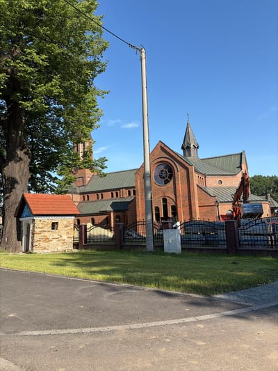 The image depicts the Church of St. Hedwig in Stanowice, Poland, under what appears to be ongoing construction or maintenance. The brick church features a prominent steeple and a large circular stained-glass window above the main entrance. An orange excavator is positioned in front of the church, its bucket raised, suggesting recent activity. Several stacks of building materials, possibly bricks or pavers, are visible behind a black metal fence surrounding the church grounds. To the left of the church, a small, rustic stone structure with a red-tiled roof stands near a large, mature tree. The scene is illuminated by bright sunlight on a clear blue sky. A concrete utility pole with electrical wires runs vertically through the left side of the frame. The area in the foreground shows an asphalt road with a light-colored stripe.