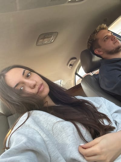 The scene depicts the interior of a passenger vehicle, identified as being in Golyamo Belovo, Bulgaria. Two individuals are present.
In the foreground, occupying the front passenger seat, is a female subject with long, dark brown hair parted on the left, cascading over her left shoulder and chest. She is wearing a light grey crew-neck sweatshirt. Her left hand, with light-colored nail polish, rests near her chest. She faces directly towards the camera, with a neutral expression.
In the background, to the right, is a male subject occupying the driver's seat. He has short, spiky, light-colored hair and visible facial stubble. He is wearing a dark-colored hooded garment. His head is turned upwards and to the right, directing his gaze outside the vehicle's frame.
The vehicle's interior features a light beige headliner with a clear plastic dome light fixture centrally located. A beige grab handle is visible above the passenger-side door. Dark-colored car seats with headrests are present. In the background, behind the passenger seat, a brown cardboard box with the partial inscription "Airtac" is discernible. Portions of the vehicle's windows reveal diffuse daylight and hints of an exterior building structure. The female subject appears to be taking a photograph. No direct interaction between the subjects is evident.