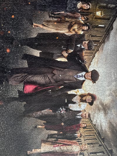 The image is a photograph of a jigsaw puzzle depicting a group of people in vintage clothing. The scene appears to be set in a city at night, with a lamppost visible in the background and some orange glowing embers on the ground. The people are arranged in a line, some looking at the camera and others looking away. They are all dressed in suits, hats, and dresses that suggest a historical period, possibly the early 20th century. One person in the foreground is holding a cigarette. The puzzle itself is laid out on a surface, with its pieces clearly visible. The image does not contain any information about the city of Craigavon, United Kingdom.
