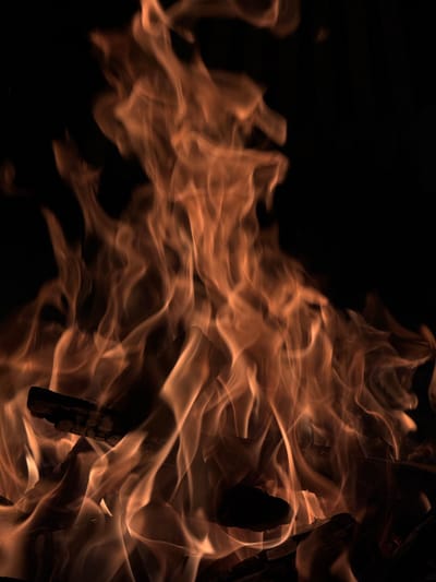 A close-up, vertical shot captures a vibrant fire burning against a dark background. The flames, in shades of orange and yellow, twist and curl upwards, with occasional hints of red. Visible within the fire are charred pieces of wood, suggesting a campfire or bonfire. The image is devoid of any human presence, and the environment is obscured by the darkness and the intensity of the flames. The setting does not provide specific indicators of its location within Vardablur, Armenia, other than what might be inferred from general rural or outdoor contexts associated with fires.