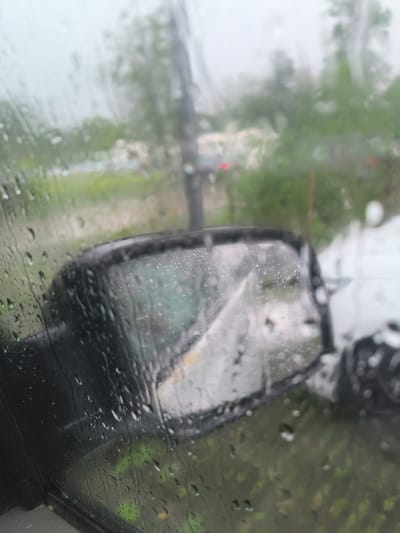 The image captures a close-up view from inside a vehicle, looking out through a side window heavily streaked and beaded with raindrops. The focal point is the dark side mirror, also wet with rain, reflecting a blurry image that appears to be a road or lane with some indistinct shapes, possibly another vehicle, in the distance. The background beyond the window and the mirror is significantly blurred, depicting a hazy outdoor scene with green foliage and hints of distant structures or vehicles. The atmospheric conditions clearly indicate rain, suggesting an overcast day. No visible text is present. The scene is set in Bad Bentheim, Germany.