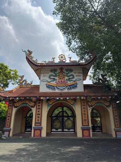 The image depicts the entrance to a Buddhist temple, identified by signage as "DRUK KARUNA MANDALA", located in Việt Trì, Vietnam. The temple features ornate architecture with a curved roof adorned with dragon figures and a dharma wheel. The facade is painted in vibrant colors, with intricate carvings and Buddhist symbols. Multiple arched gateways lead into the temple grounds, secured by decorative iron gates. Red lanterns hang from the arches, adding to the festive atmosphere. The ground in front of the temple is paved with stone tiles, and surrounding trees with lush green foliage are visible. The sky is partly cloudy, indicating it is daytime. There are no people visible in the image.