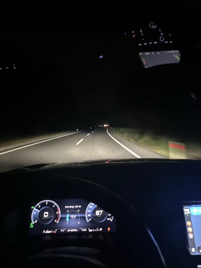 The view from inside a car driving on a dark road in Ula, Turkey. The car's headlights illuminate the road ahead, showing white lines and glimpses of the roadside. Inside the car, the dashboard is visible, displaying a speedometer reading of 87 km/h and a drive range of 269 km. A navigation system is also visible on a screen in the upper right of the frame.
