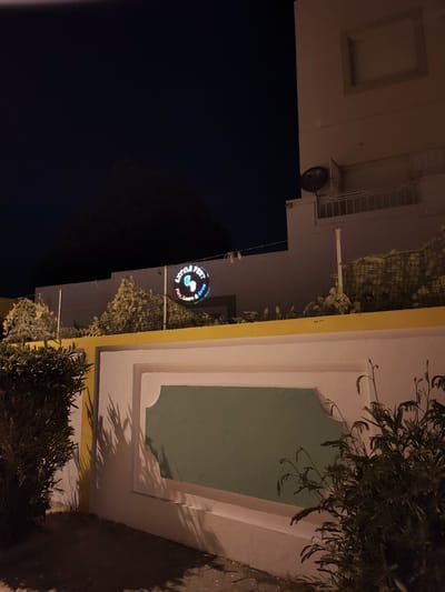 An outdoor, low-angle, night-time, wide shot captures a building with a sign illuminated by lights. The sign, which is circular, displays the words "Little Feet" with an image of feet. Below this, it reads "Play, Learn & Grow". The building is light-colored with a yellow band across the top of a lower wall. Green foliage is visible in front of the building, casting shadows on the wall. A darker building is seen to the right of the frame, with a satellite dish mounted on its side. The sky is dark.