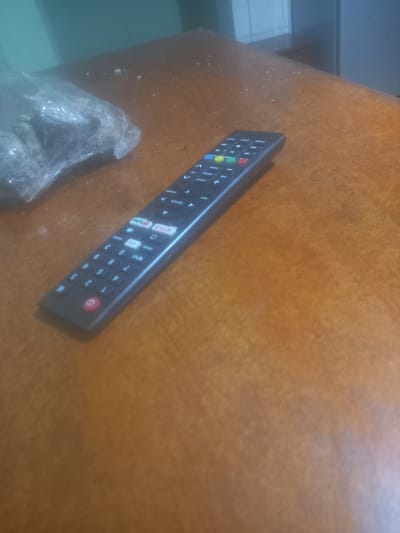 A black TV remote control lies on a brown wooden table. To the left of the remote, there is a clear plastic bag containing some dark, granular material. The scene appears to be indoors, with a wall in the background that is partially visible and appears to be painted a teal color. The lighting in the room is dim.