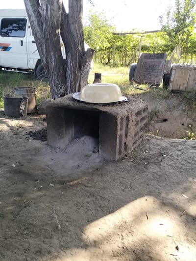 In Aktam (Tashminvody), Uzbekistan, an outdoor scene unfolds on a dry, dusty ground, illuminated by sunlight casting distinct shadows. A primitive clay oven, constructed from bricks and mud, stands prominently in the foreground. It has an opening at the front, revealing ash and remnants of previous fires. A large, pale yellow metal bowl rests on top of the oven. To the left of the oven are two large, weathered metal buckets, one filled with dry, straw-like material. Behind the oven, a large tree with rough, textured bark dominates the scene. In the background, a white van with orange and blue stripes is parked, and beyond that, a vineyard with trellises and lush green foliage is visible. Several wooden crates and a rusty, two-wheeled trailer are also present in the mid-ground. The overall environment suggests a rural or semi-rural setting, possibly a backyard or communal area.